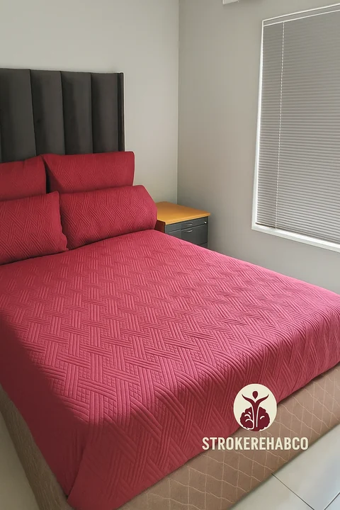 Made bed with a burgundy quilt and pillows, dark upholstered headboard, bedside cabinet and window blinds; StrokeRehabCo branding appears at the bottom of the photo.